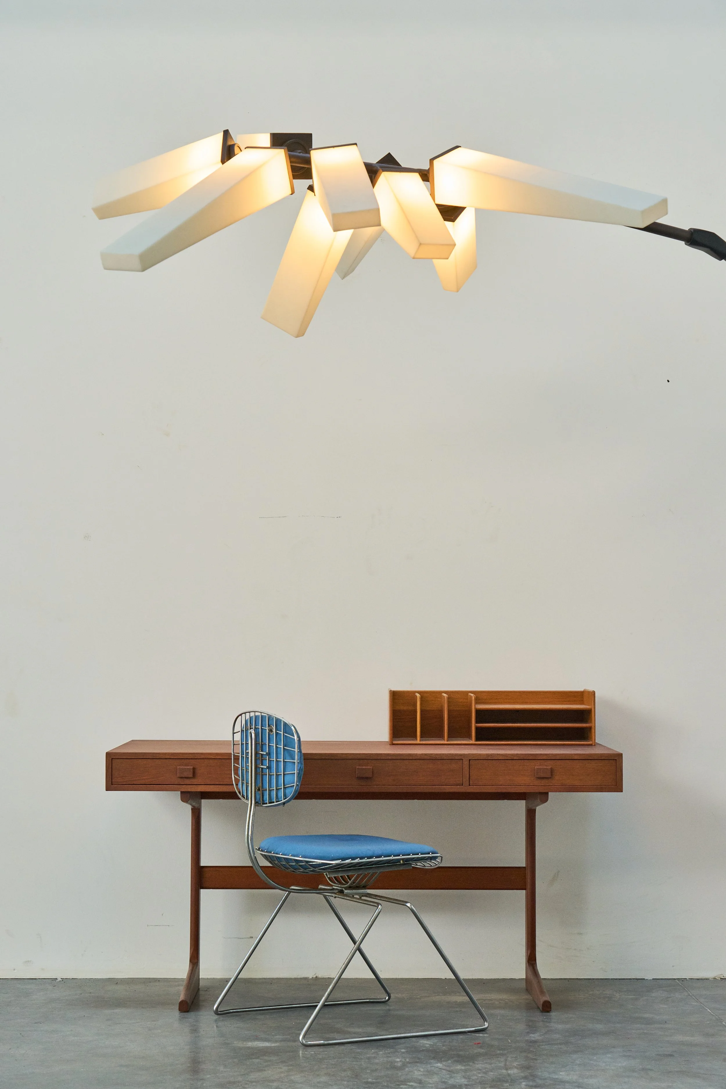 An interior scene with a minimalist wooden desk and a vintage-style wire chair with a blue cushioned seat. Above the desk is a modern chandelier with shaped white light fixtures hanging from a black mount. The background features a plain white wall.