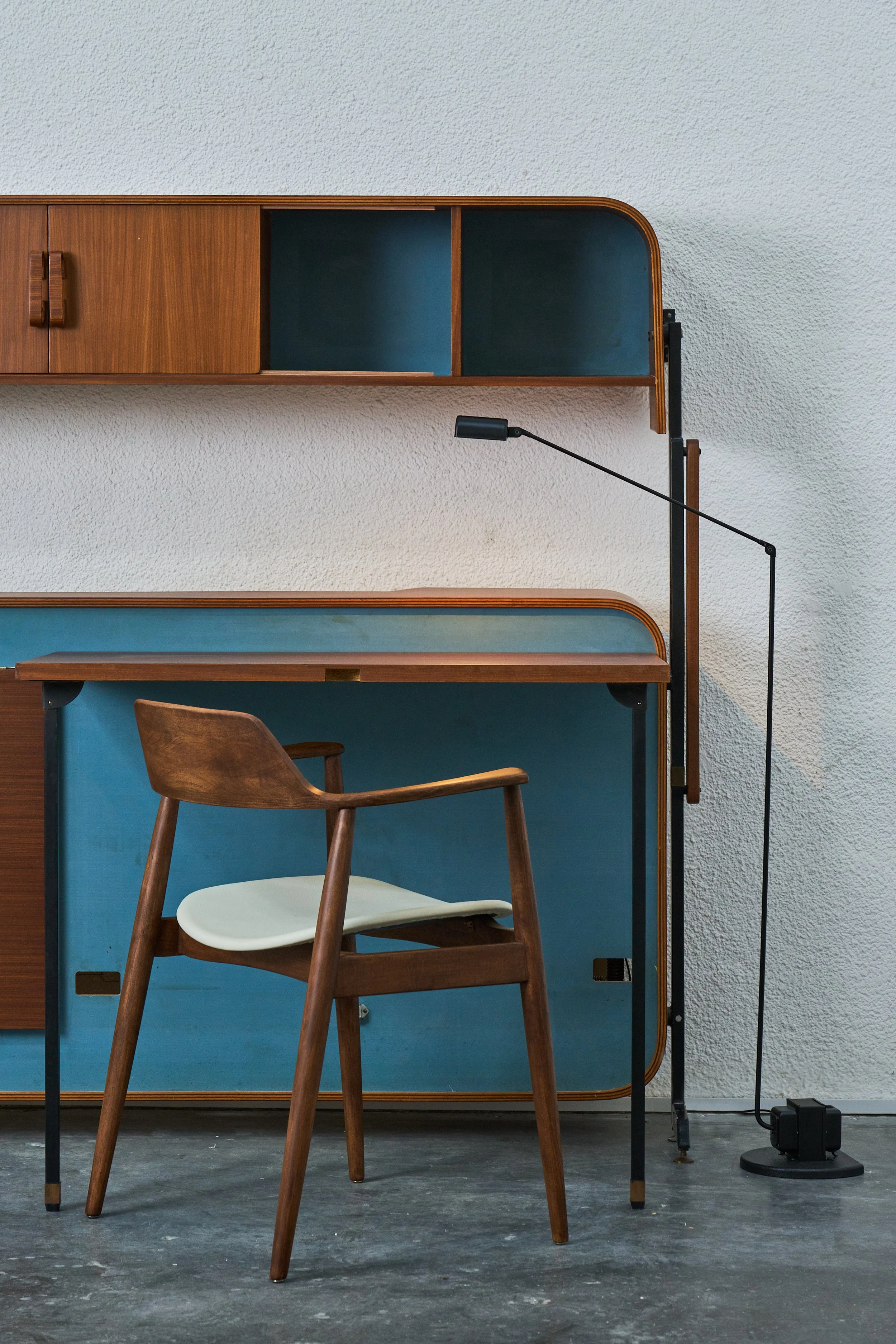 A wooden chair with a white cushion in front of a vintage blue and wood desk, with a black adjustable desk lamp on the side, set against a textured white wall and a gray concrete floor.