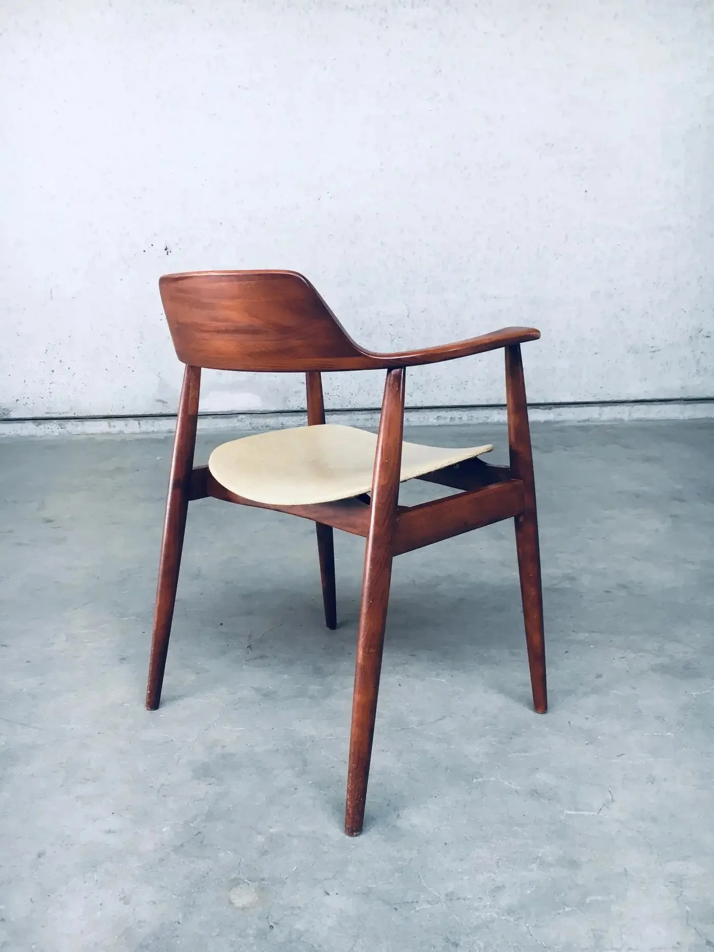A wooden chair with a curved back and a light-colored seat, placed on a concrete floor against a plain white wall.