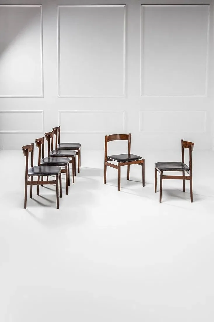 Six wooden chairs with black seats arranged in a semi-circle against a white wall.