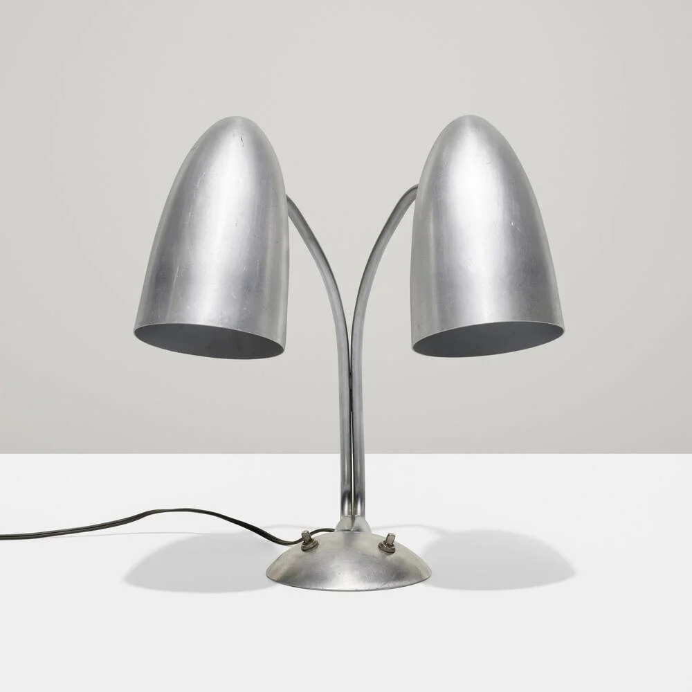 A modern silver desk lamp with two adjustable conical lampshades, placed on a white surface against a light background.