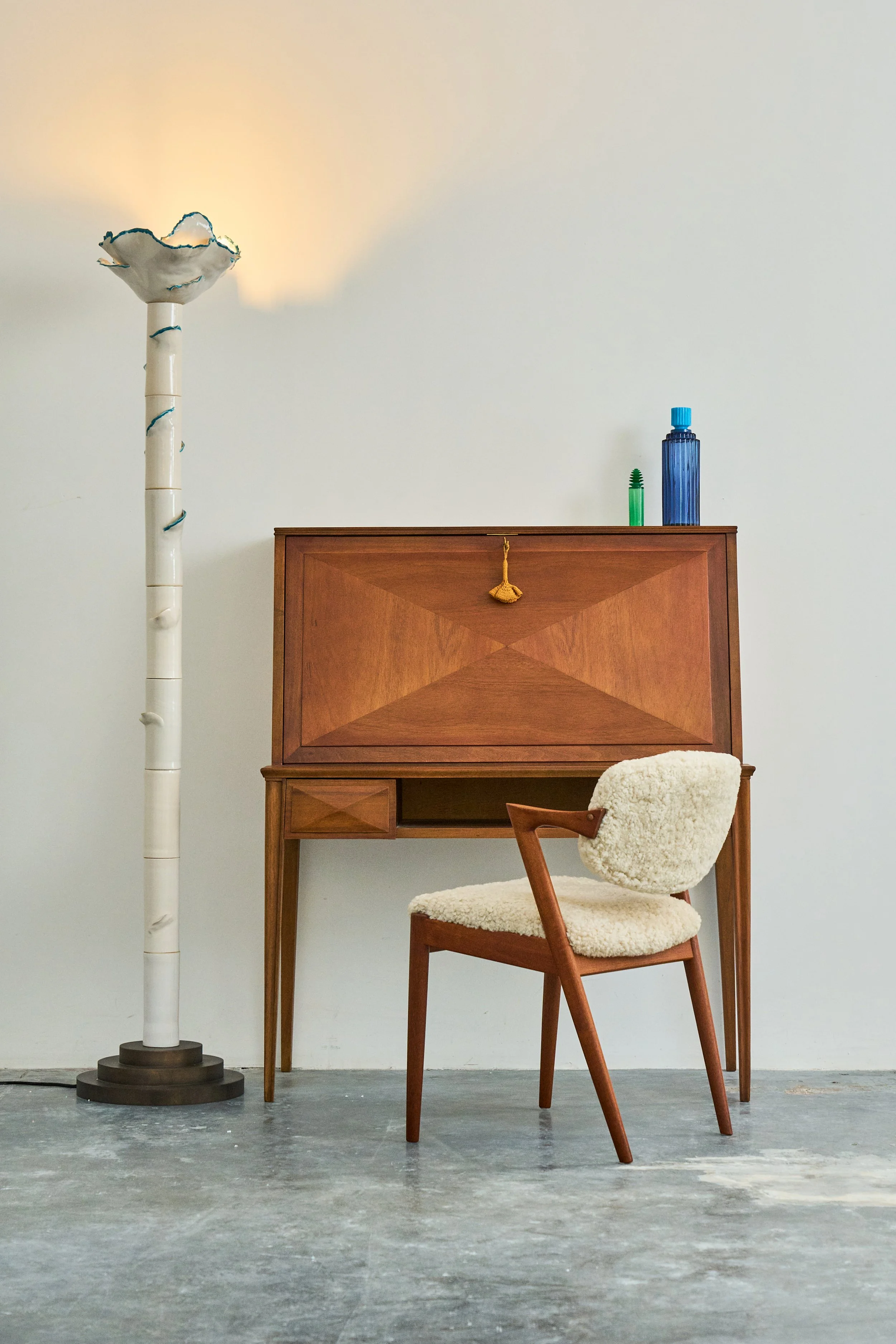 A wooden desk with a vintage style chair with a fluffy white cushion, a tall white floor lamp with aflower-shaped shade, two decorative glass bottles on the desk, and a plain white wall background.