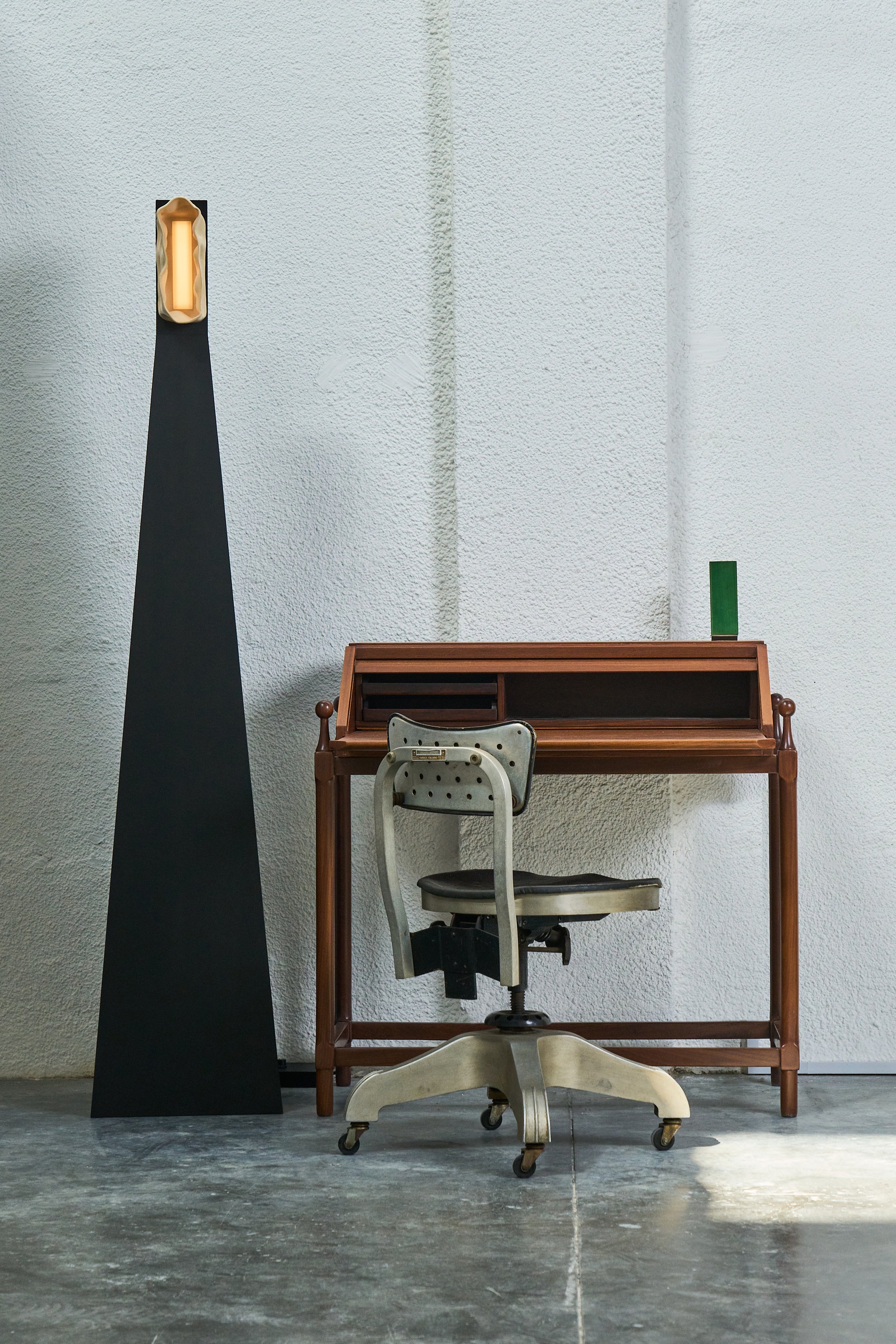 A vintage office or school desk with a rolling chair in front of it, placed against a textured white wall. There are two objects on top: a green container on the right side of the desk and a tall, black triangular sculpture with a glowing vertical light on the left side. The scene has an industrial and minimalist aesthetic.