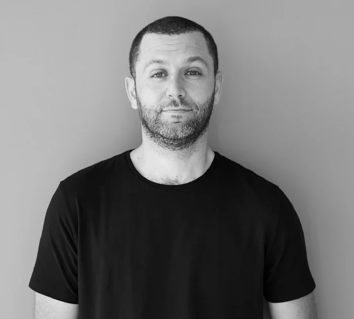 Black and white portrait of a man with short dark hair, a beard, and a slight smile, wearing a plain black t-shirt, standing against a plain background.