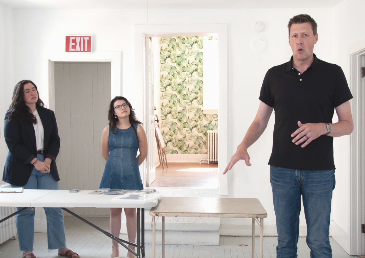 A man appears to be speaking or explaining something in front of two women during a meeting or presentation in a room with white walls and a doorway with tropical leaf wallpaper in the background.