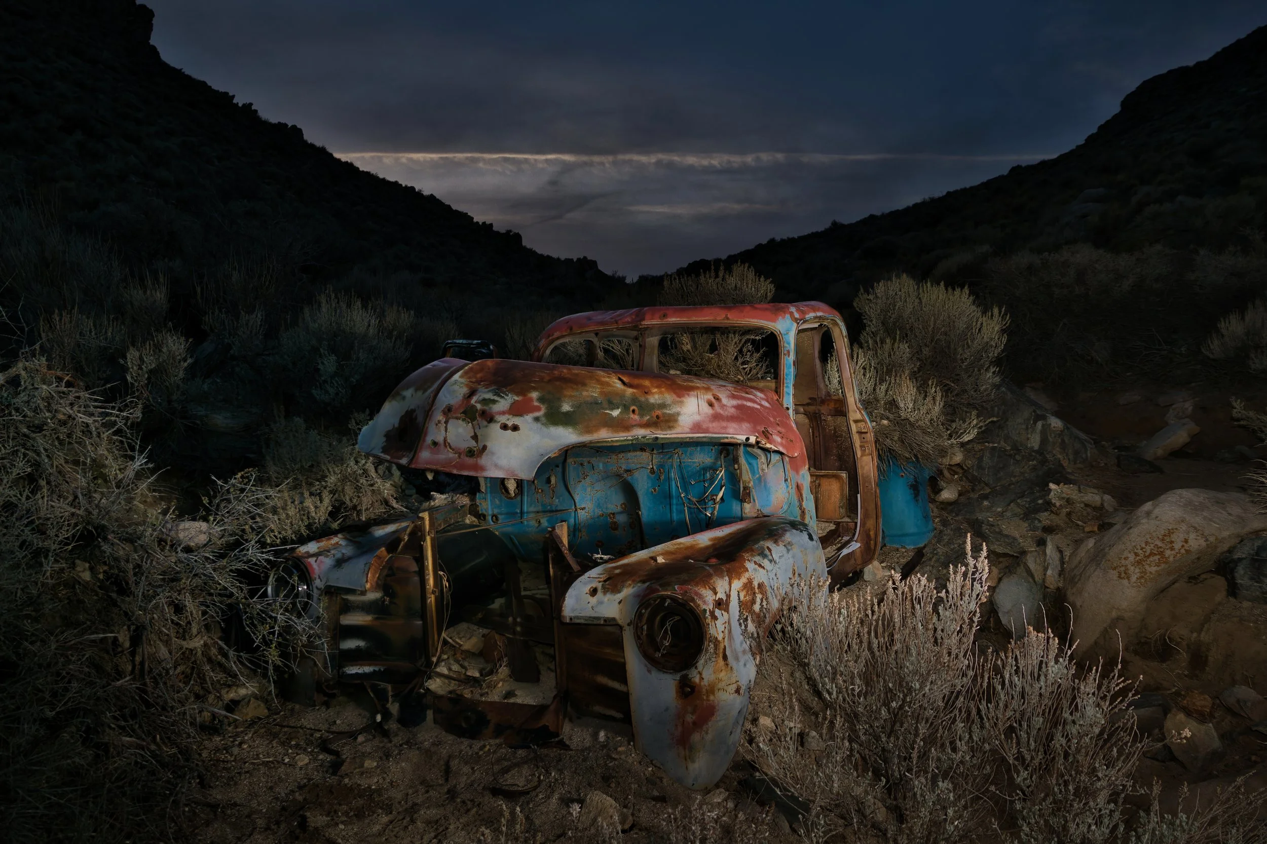 Rusty, abandoned truck in a desert landscape during dusk with mountains and dark sky in the background.