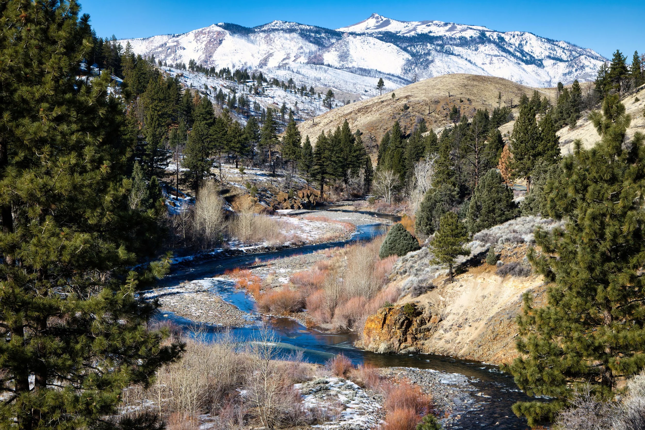 A scenic landscape of a winding river flowing through a mountainous terrain with snow-capped peaks in the background, surrounded by evergreen trees and dry shrubs.