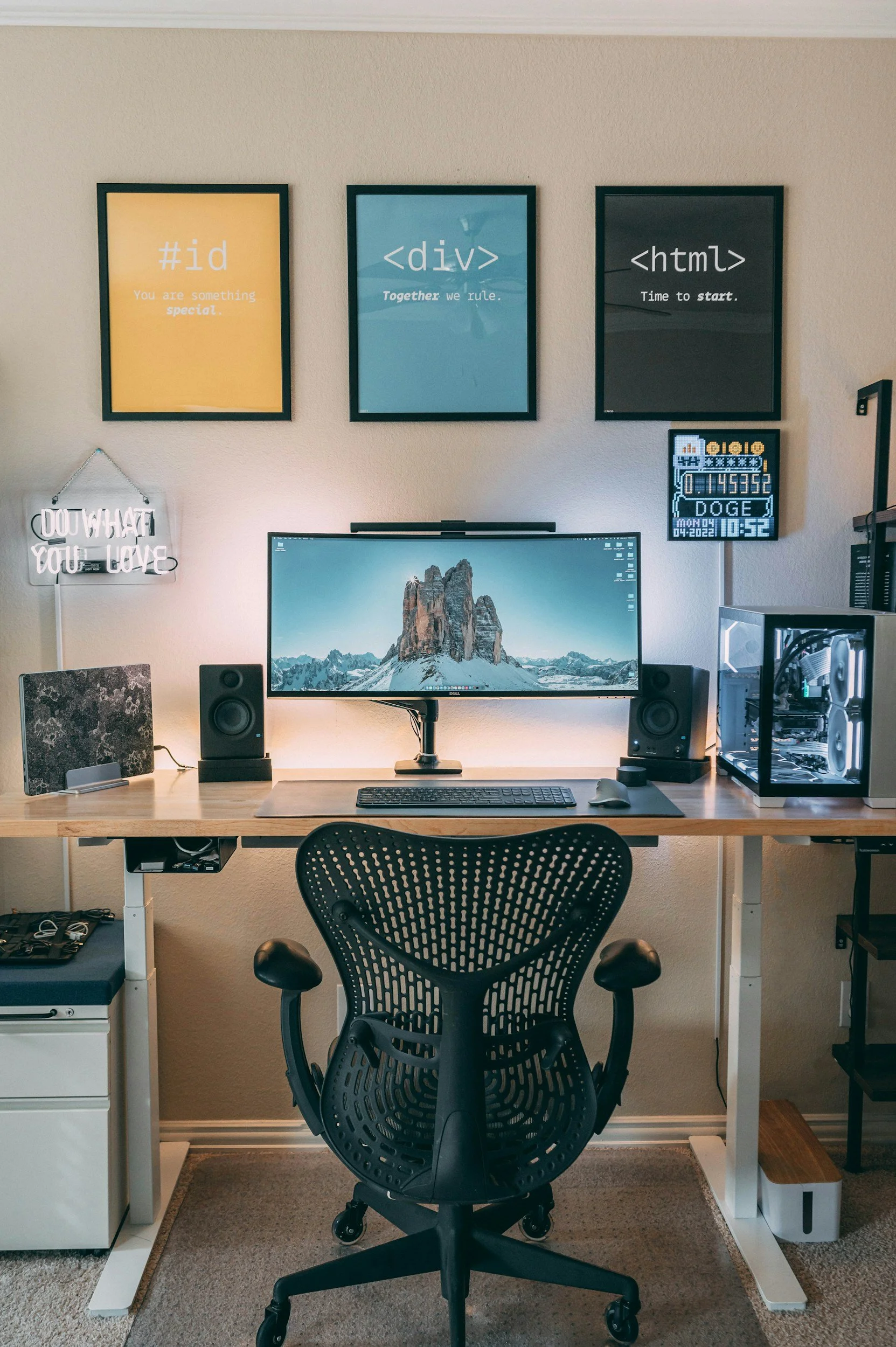 A modern computer desk setup with a large monitor, keyboard, mouse, speaker, and a computer tower with visible internals. Above the desk, three framed prints display coding symbols and phrases, and there is a neon sign on the wall to the left. A digital clock displaying the time, date, and weather information is also present.