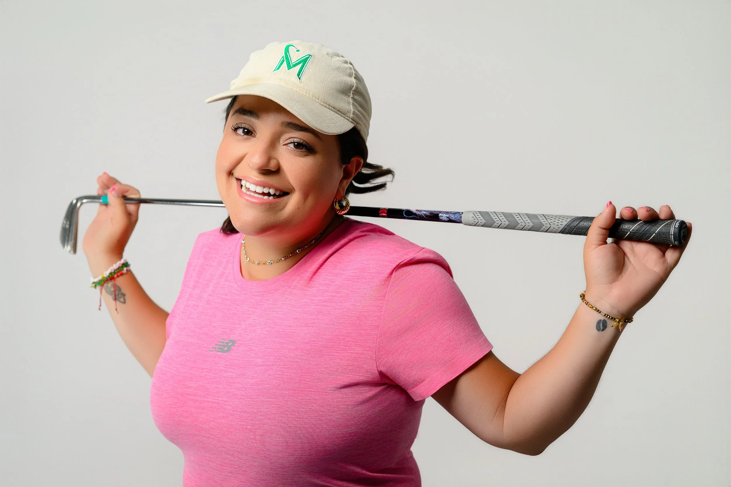 A smiling woman in a pink t-shirt and beige cap holding a golf club over her shoulders against a plain white background.