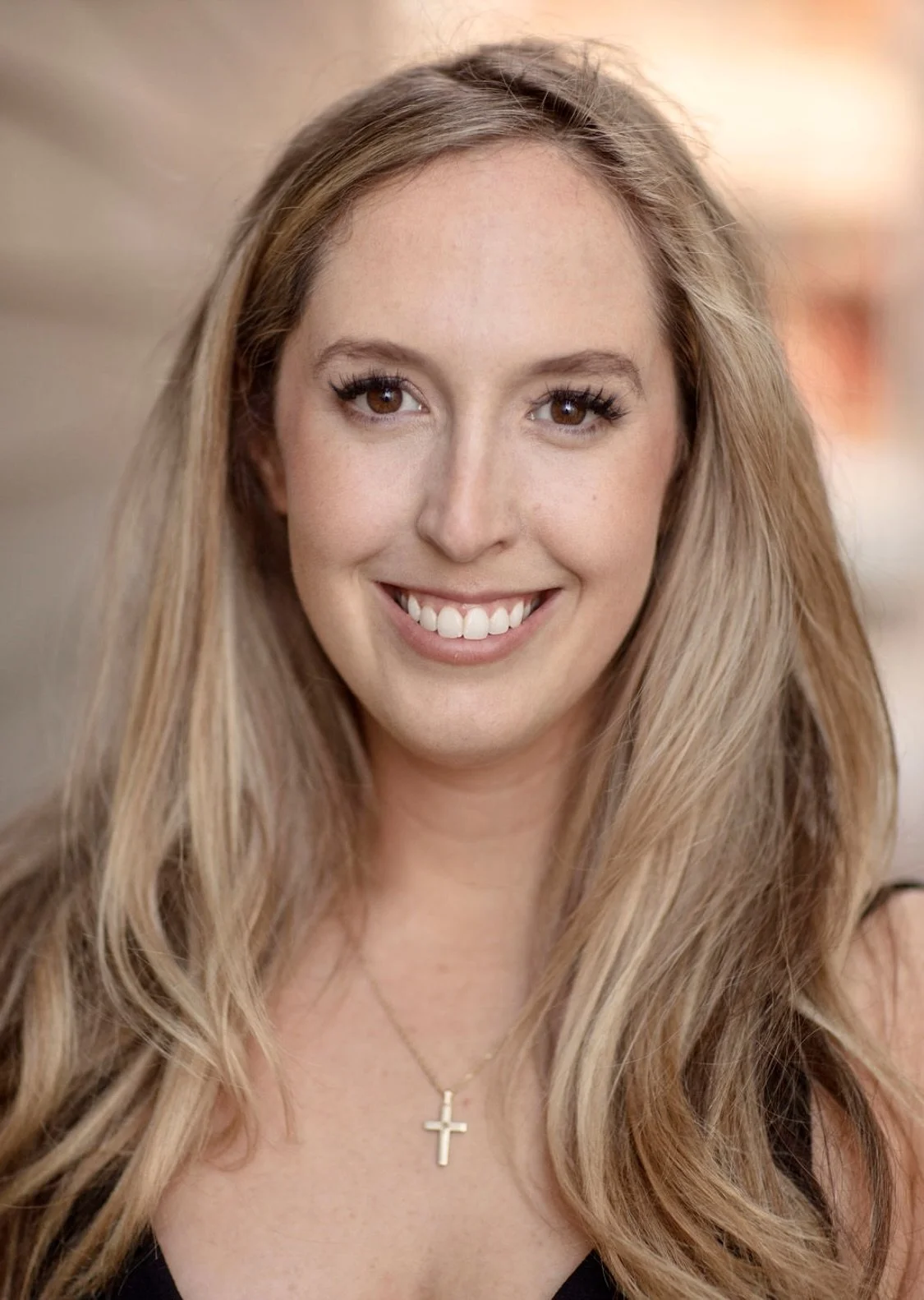 A smiling woman with long blonde hair, wearing a necklace with a cross pendant, in an indoor setting with warm background lighting.