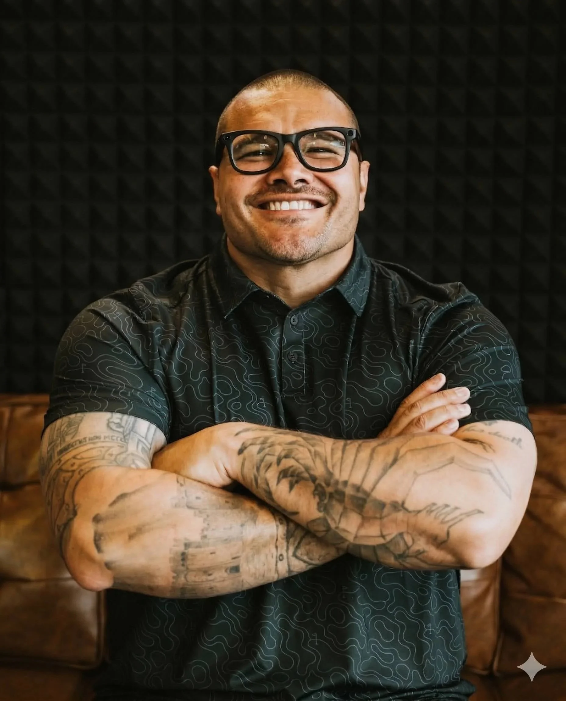 A smiling man with glasses and tattoos on his arms, wearing a black shirt with a subtle pattern, sitting with his arms crossed in front of a dark, textured background.