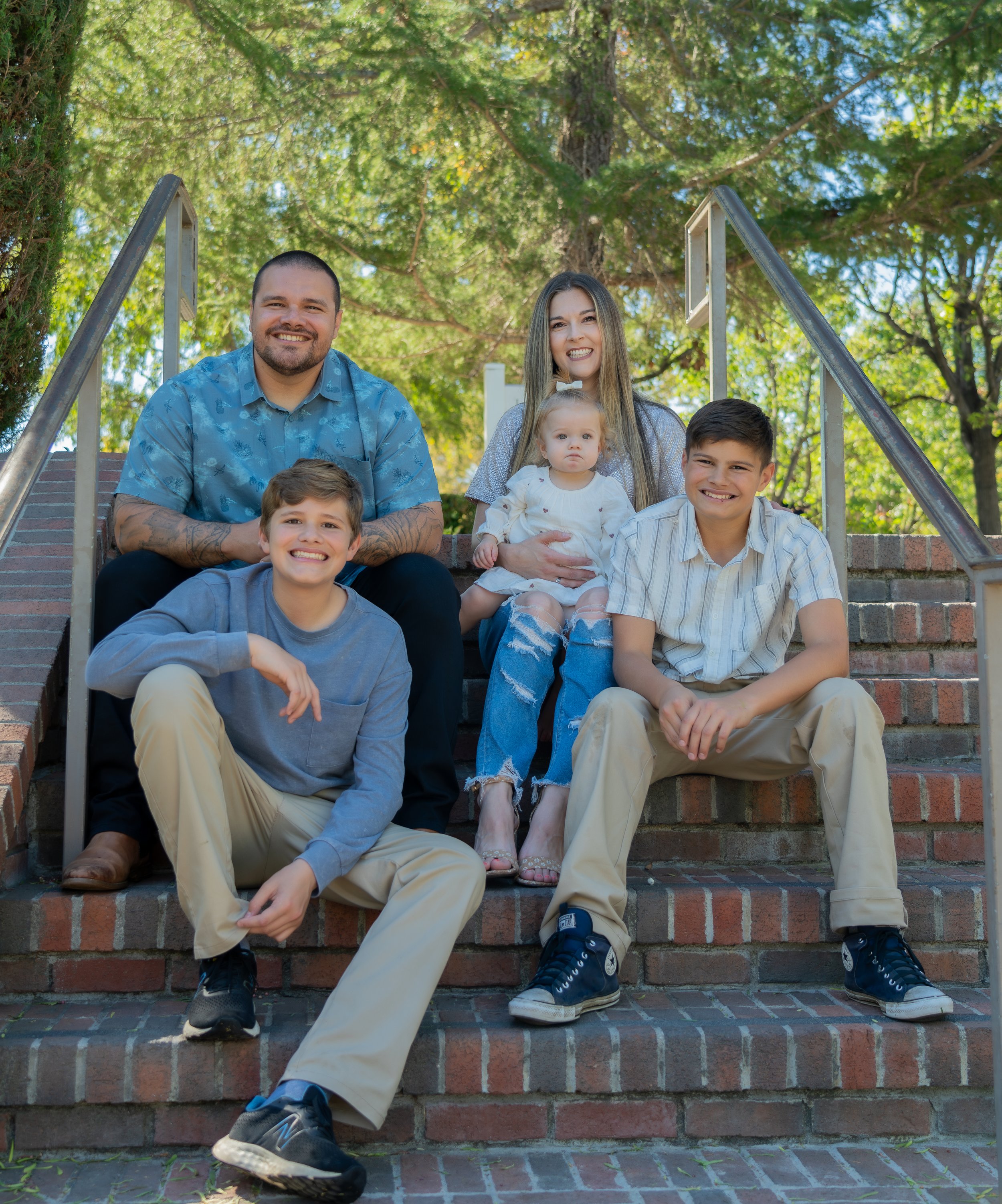 A family of six sitting on brick stairs outdoors, smiling and posing for a photo. The group includes two adult men and women, a teenage boy, a young boy, and a young girl. The background features green trees and sunlight.