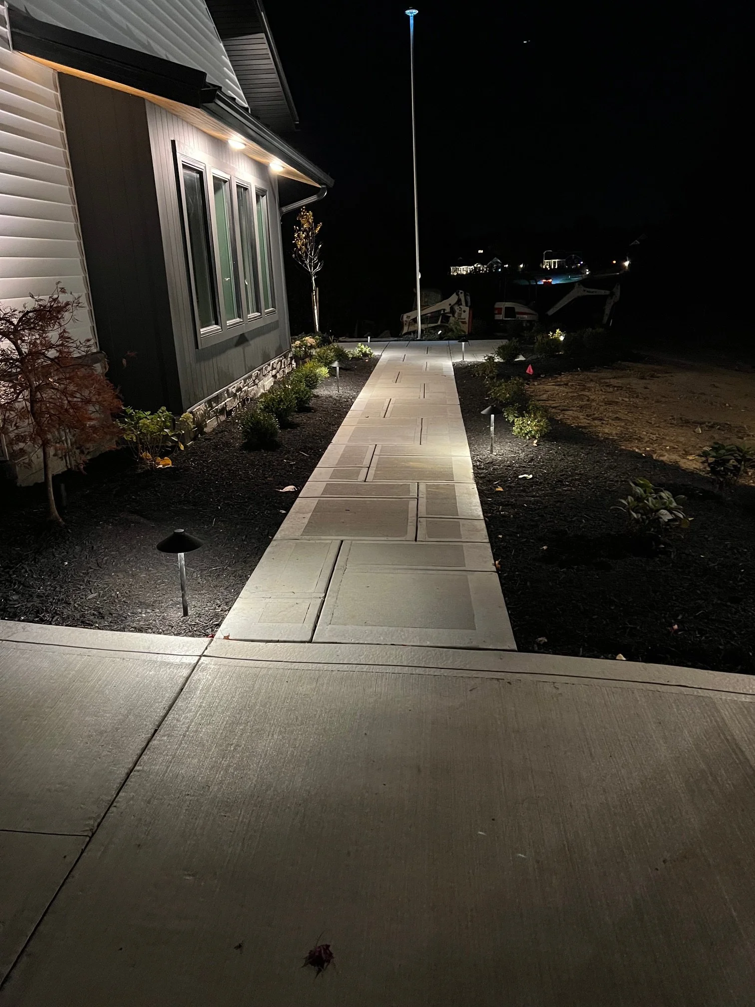 Nighttime view of a well-lit concrete walkway leading to the house, with small landscaped plants and garden lights on both sides, outdoor lighting for walkways done by Salupo Lawn Sprinkling