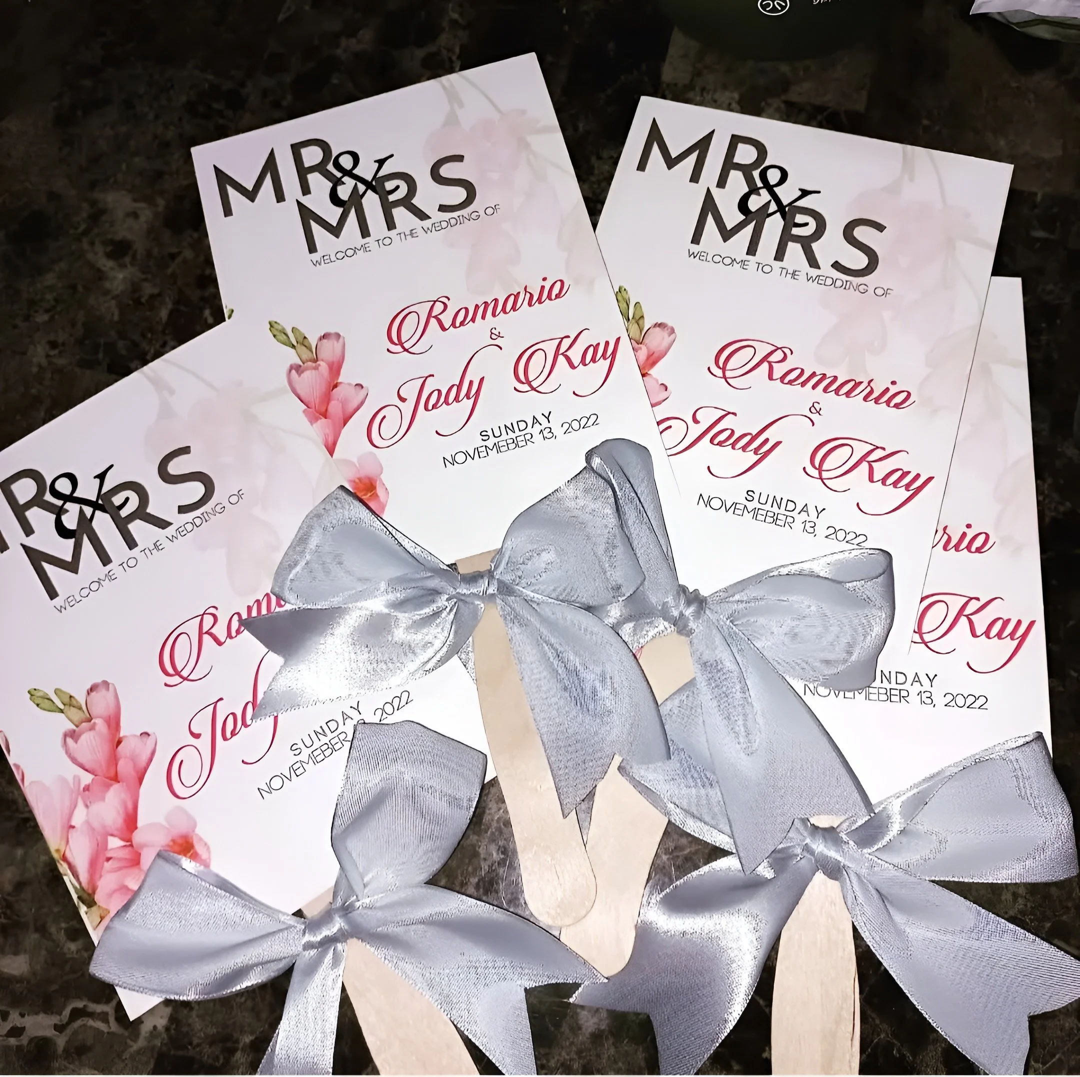 Wedding programs with pink flowers, black and red text, and silver ribbons on a dark surface.