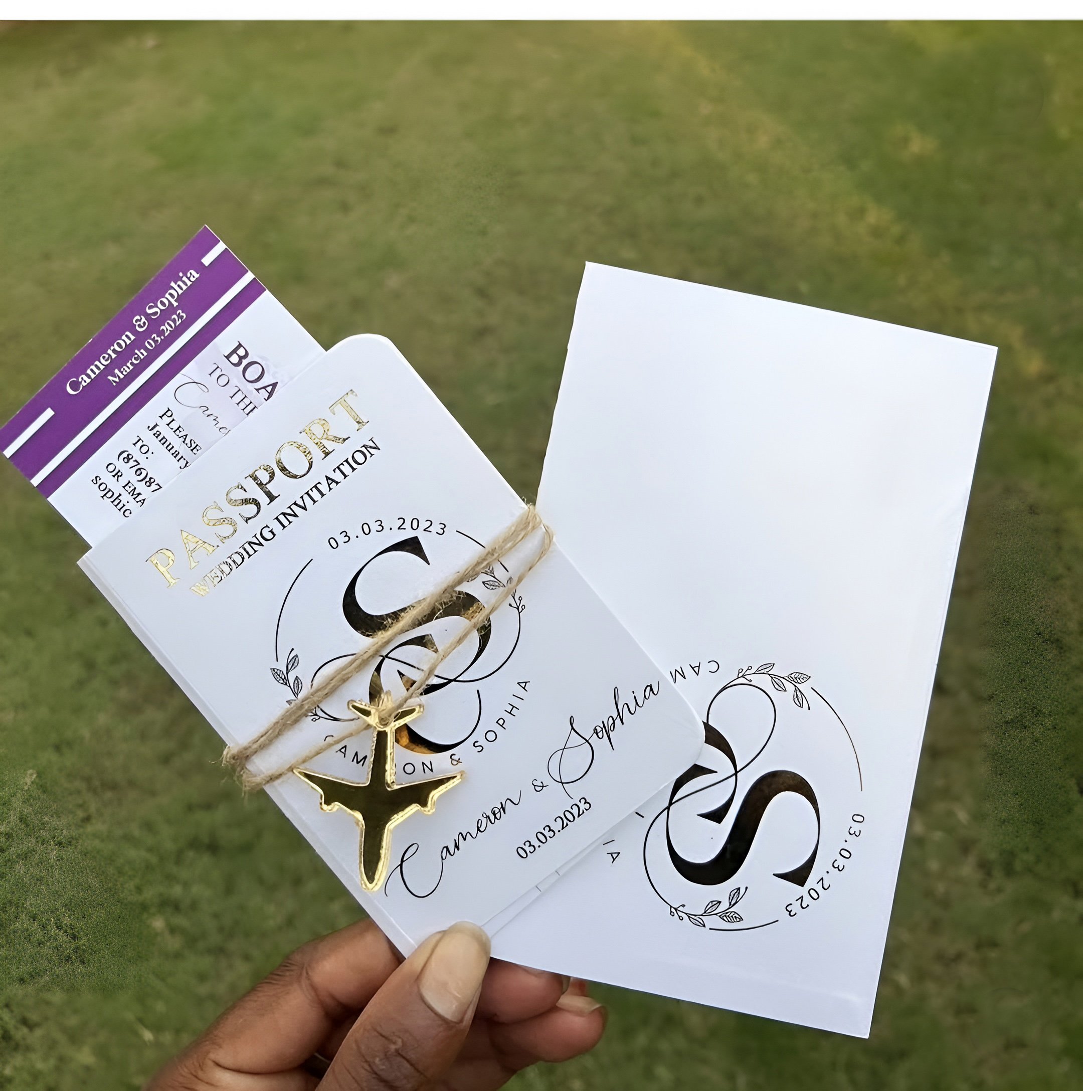 Wedding invitation suite with a white card featuring floral design and the couple's names, a purple RSVP card, and a small white card, along with a gold anchor-shaped charm attached to twine. The background is green grass.