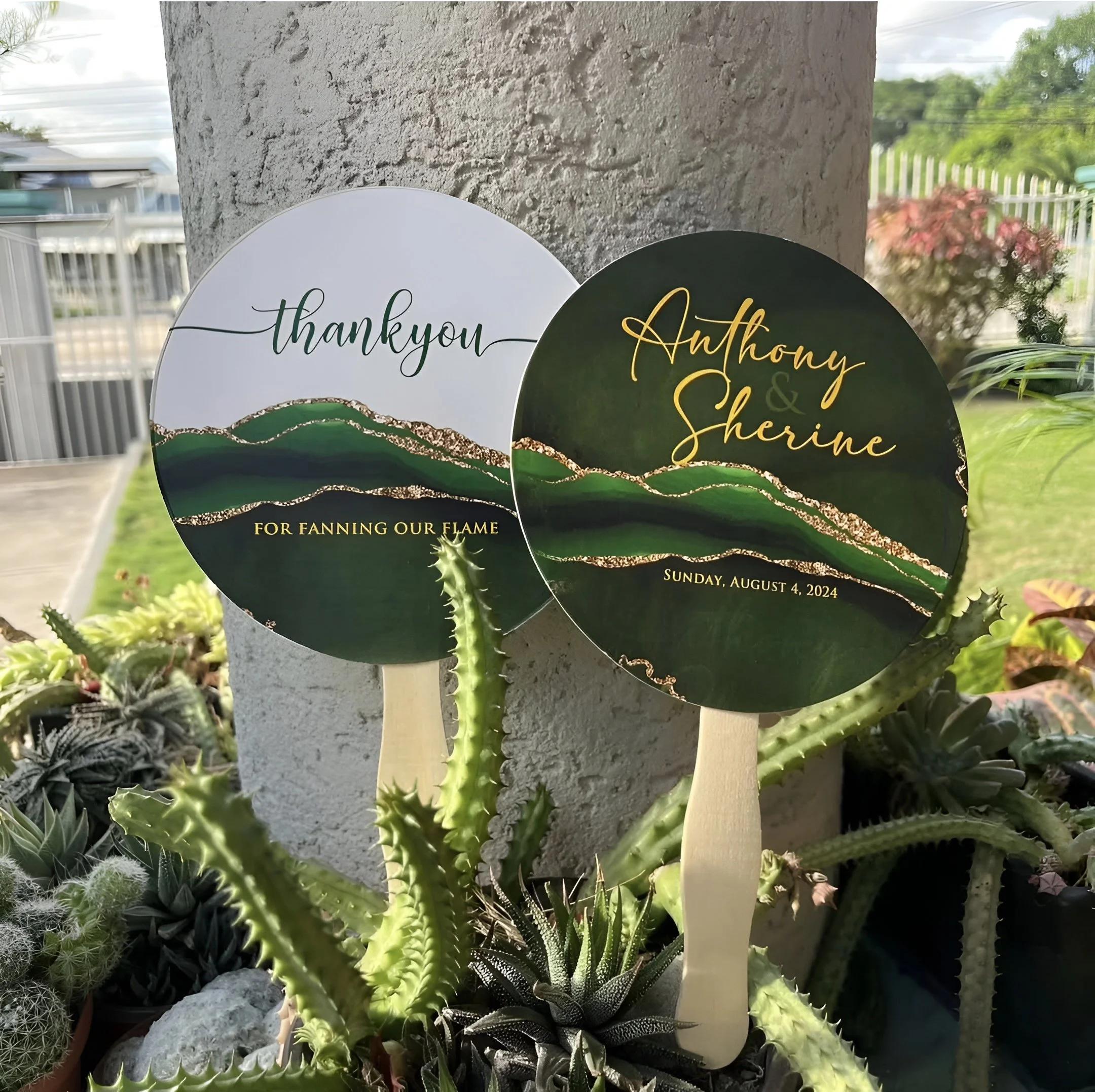 Two round wedding signs with green and black backgrounds, gold and green accents, and wooden sticks, placed among various cacti, with a textured white wall behind them and a garden in the background.