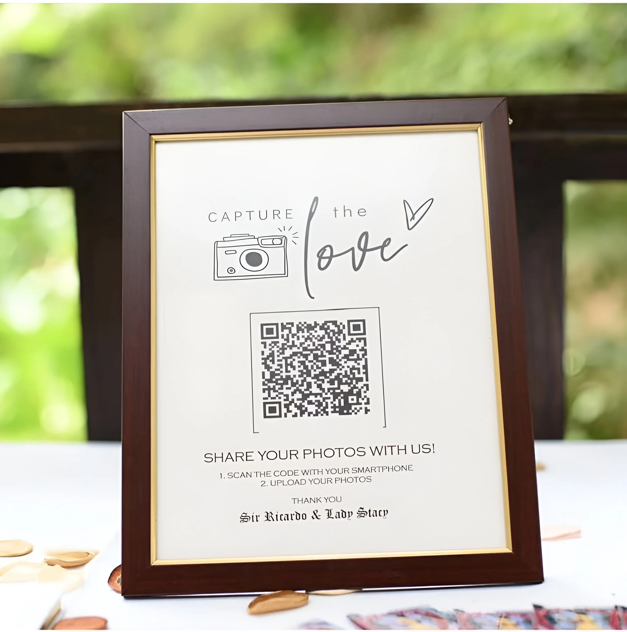 A framed sign on a table with a white background and gold and brown border, displaying instructions for sharing photos via a QR code at an event for Sir Ricardo & Lady Stacy, with a camera illustration, a heart, and handwritten-style text.