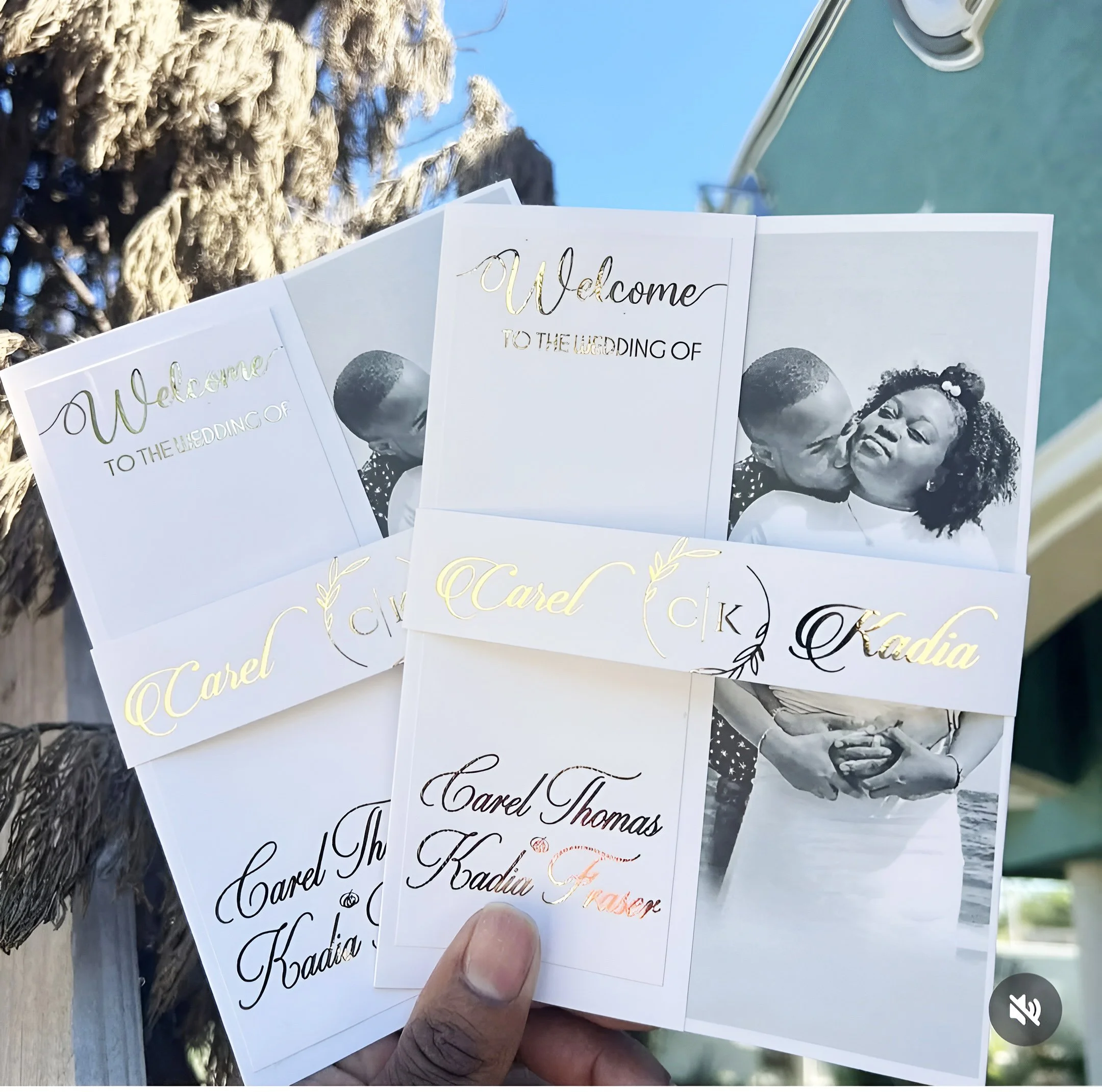 Wedding invitation cards featuring pictures of a couple and elegant gold and black script text, held by a hand outdoors against a blue sky and a green building.
