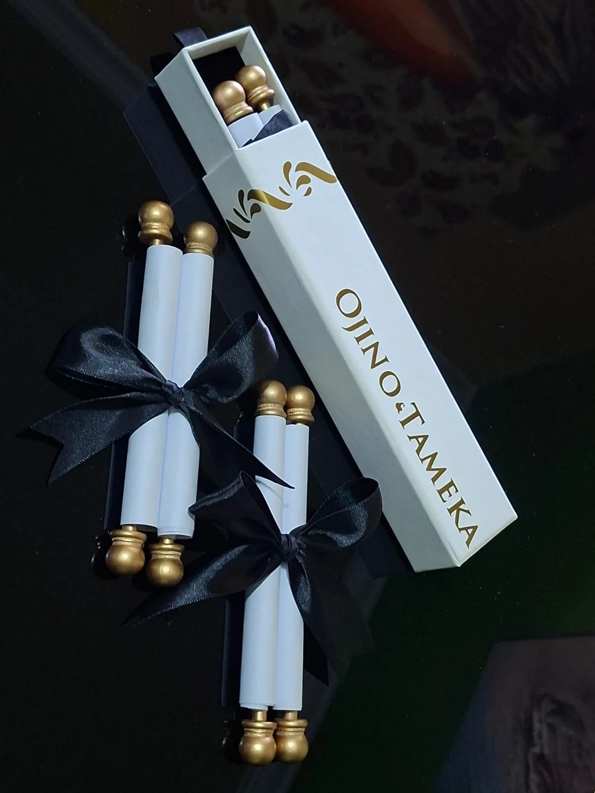 A set of white candles with gold tops, tied with black ribbons, and a white box labeled 'OONO TAM'EKA' containing more gold-topped candles.