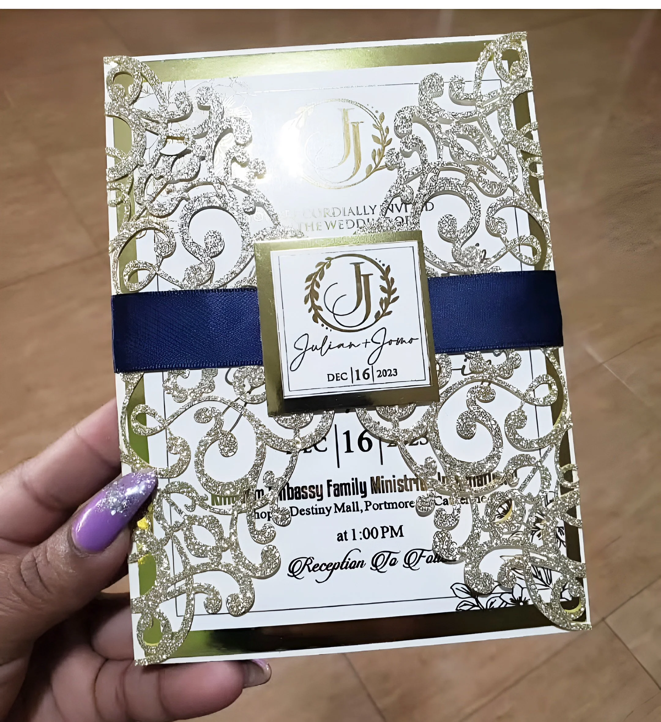 A wedding invitation card featuring intricate gold and white designs, a navy blue ribbon, and a gold-embossed monogram. The card has the names Julian and Jonna, with the wedding date December 16, 2023, and details of the reception at Destiny Mall, Po