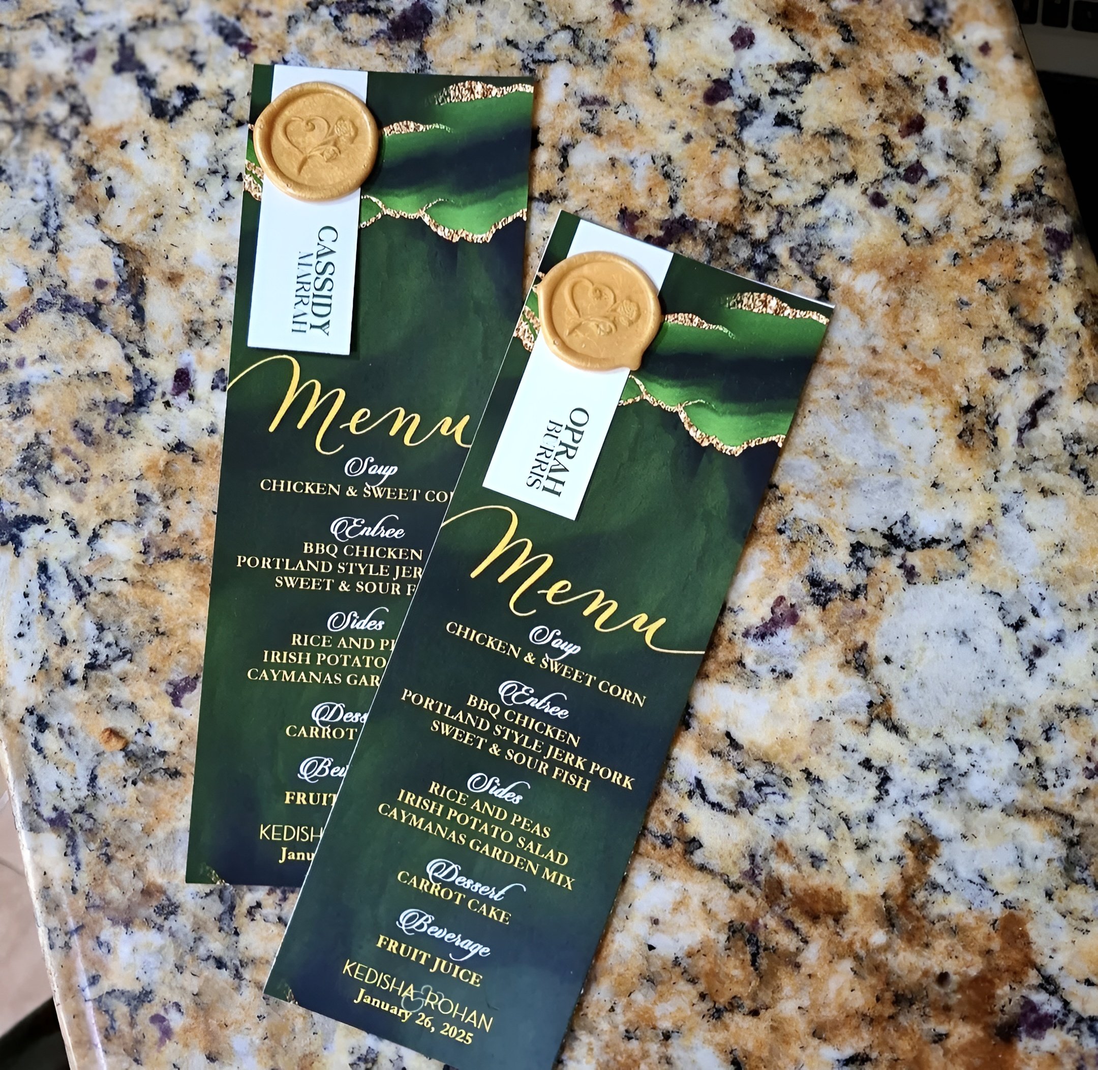 Two green and gold event menus with gold wax seals on a speckled granite countertop. The menus read 'Oprah Belize' and 'Gassidy Avari H,' with a date of January 26, 2025.