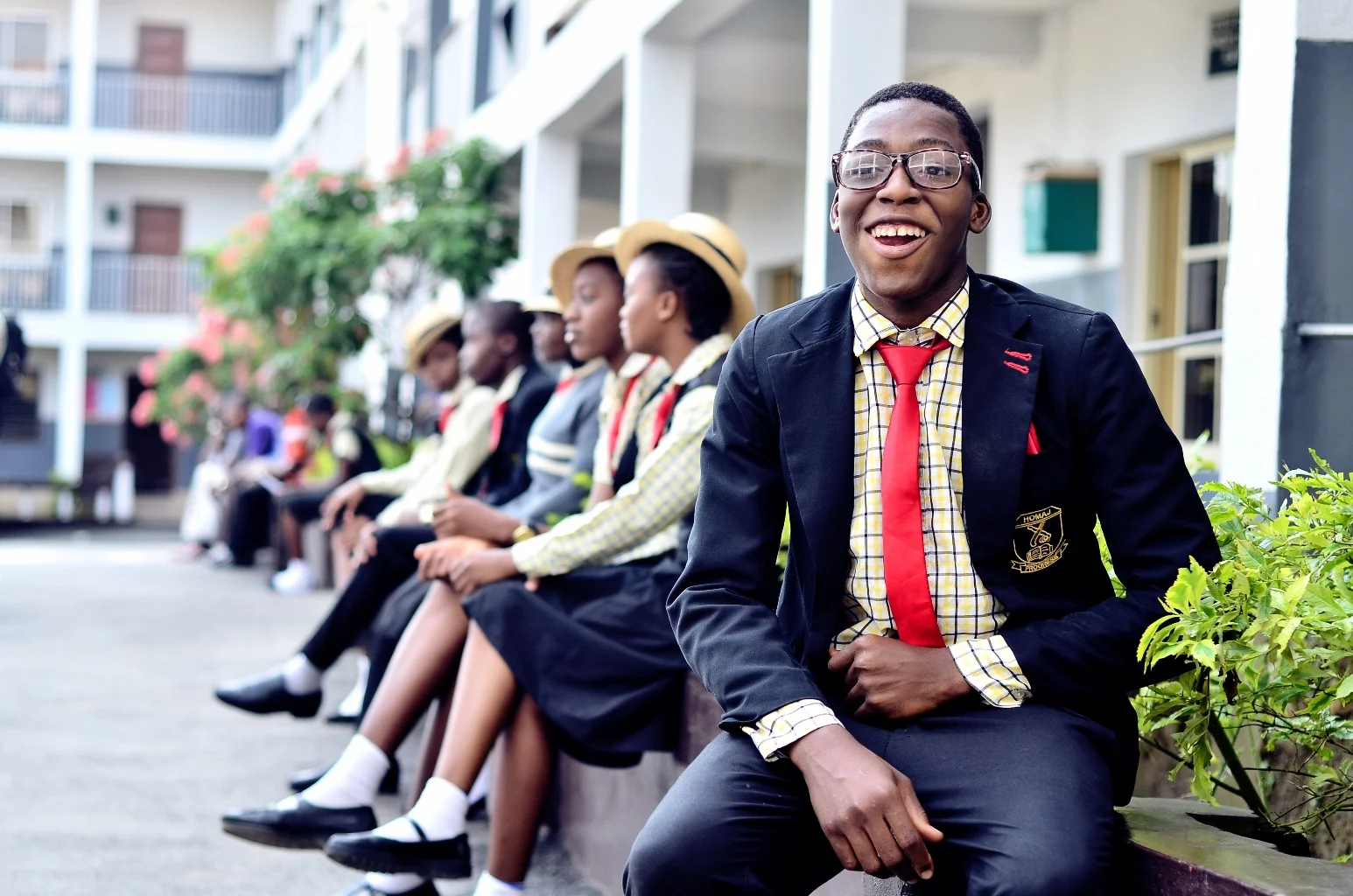 A group of  Homaj students sitting on a bench outside a school building, with one smiling student in the foreground wearing glasses, a blazer, a shirt, a tie, and a red sash.