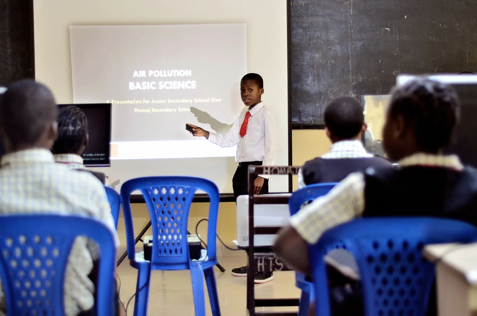A classroom scene with a young boy in a white shirt and red tie giving a presentation on air pollution to students seated at desks, with a slide on the screen behind him titled "AIR POLLUTION BASIC SCIENCE."