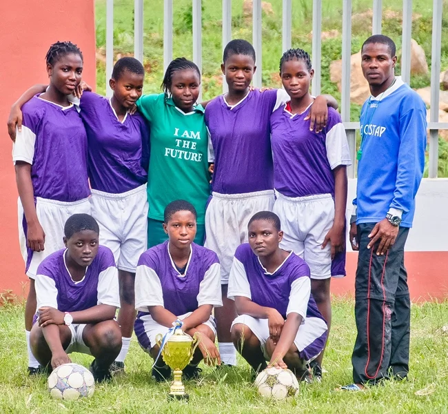Group of girls in purple and white soccer uniforms with two soccer balls and a trophy, along with a man in a blue jacket, all standing outdoors on grass.
