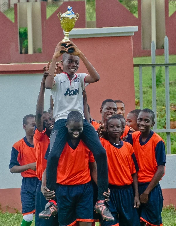 A group of young boys in orange and blue sports uniforms celebrating on a soccer field. One boy is sitting on another's shoulders, holding a trophy high above his head and smiling.