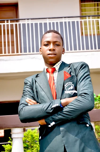 Young man in a suit and red tie standing outdoors with arms crossed, building and balcony in background.
