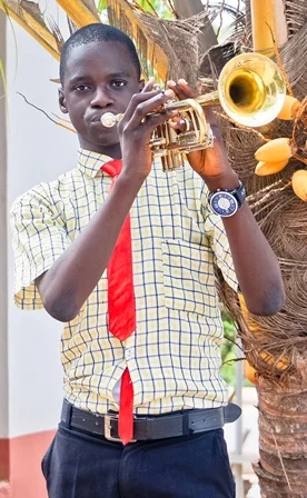 Young boy playing trumpet outdoors, wearing a checkered shirt with a red tie, and a watch on his left wrist.