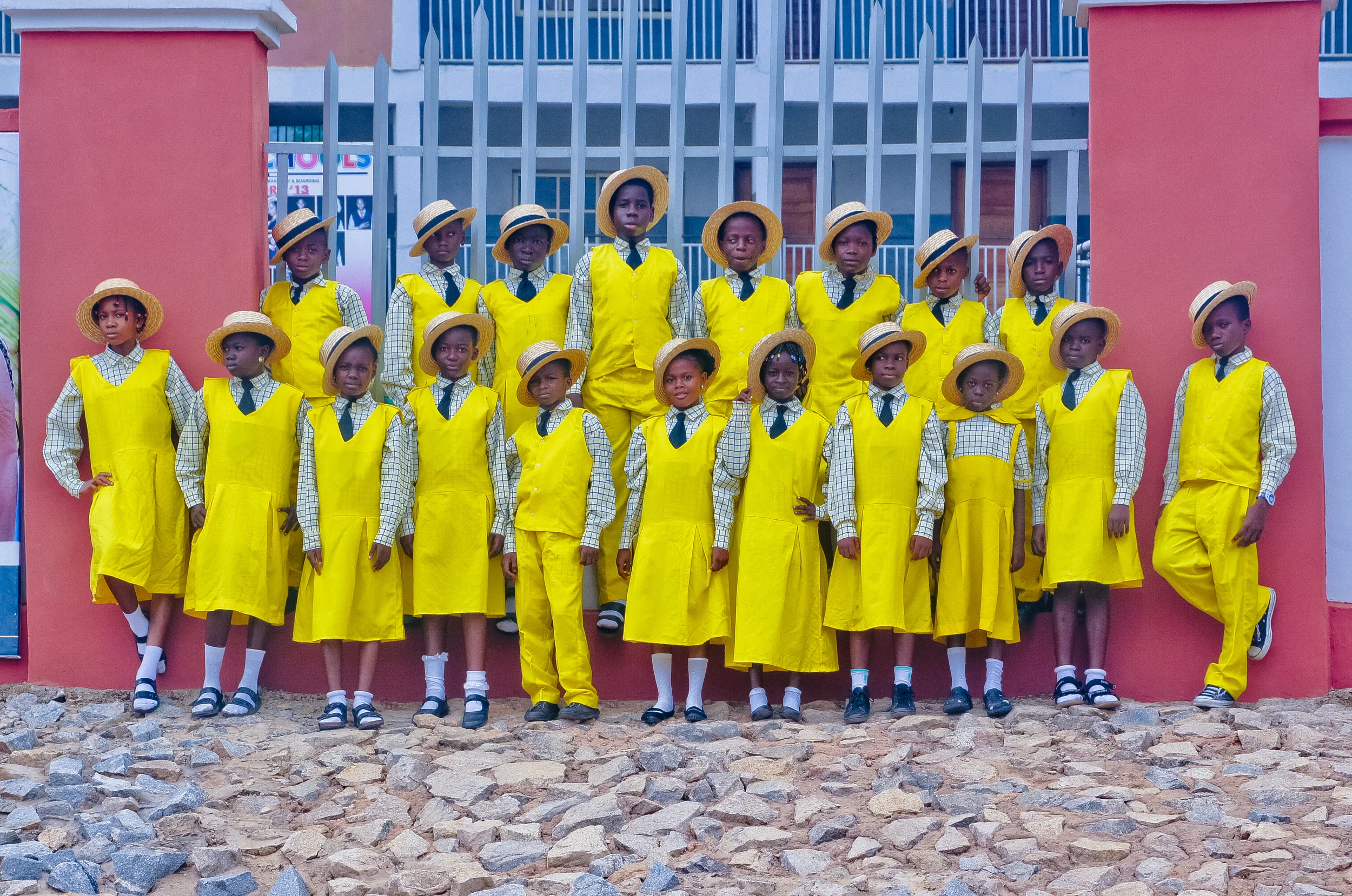 A group of school children dressed in yellow uniforms and hats, standing in front of a gate on a rocky ground.
