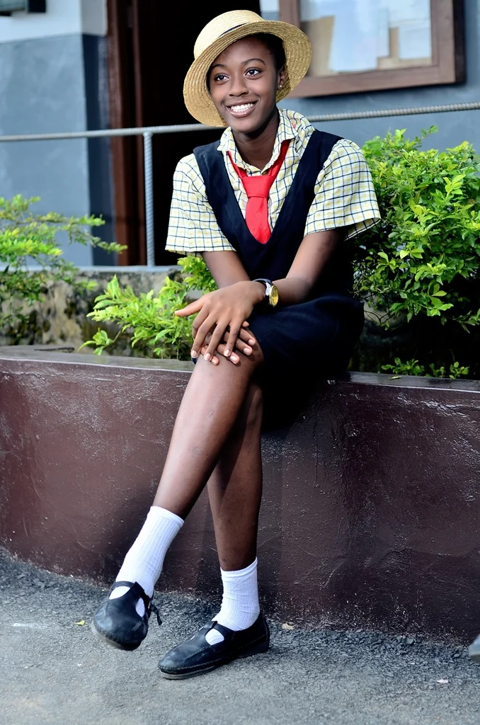 A young woman dressed in school uniform wearing a straw hat, sitting on a planter, smiling, with greenery and a building in the background.