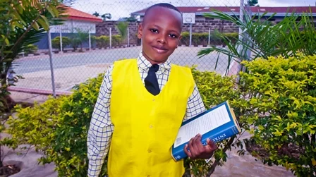 Young boy in a yellow vest and checkered shirt holding a book, standing outdoors near greenery and a fenced sports court.