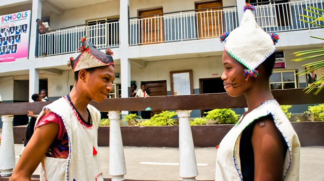 Two women wearing traditional clothing and headgear face each other on a street, with a building and people in the background.