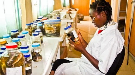 A woman in a white lab coat examining a jar in a laboratory with shelves of similar jars around her.