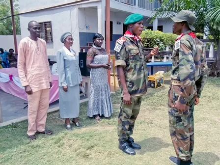 Two military personnel in uniform talking to each other outdoors, with three people standing nearby watching, in a grassy area with a building and trees in the background.