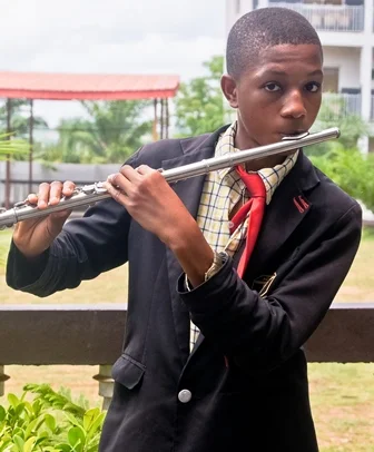 A young boy in a school uniform playing a silver flute outdoors.