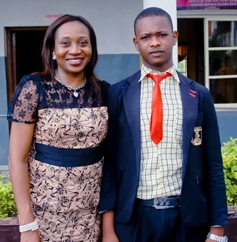 A woman in a floral dress and a young man in a school uniform standing outdoors in front of a building.