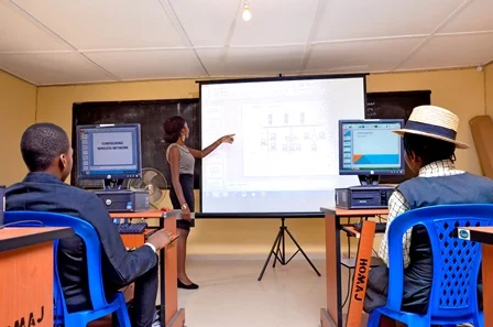 A woman stands at the front of a classroom pointing at a large projected screen, teaching three seated students using desktop computers.