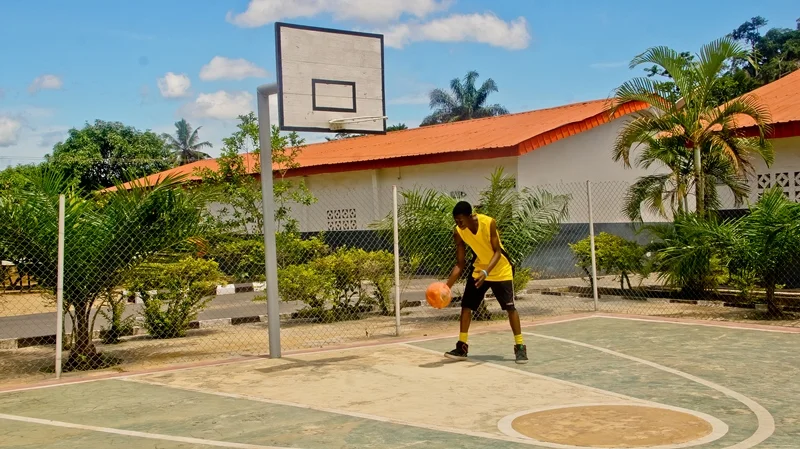 A young boy in a yellow jersey dribbling a basketball on an outdoor basketball court surrounded by a chain-link fence, tropical plants, and a building with a red-tiled roof under a blue sky.