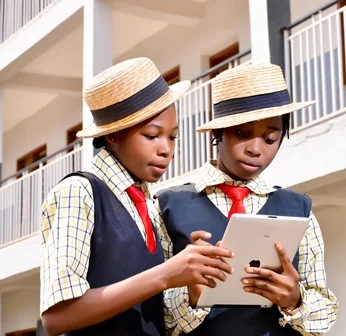 Two young girls wearing school uniforms and straw hats, looking at a tablet together outdoors.