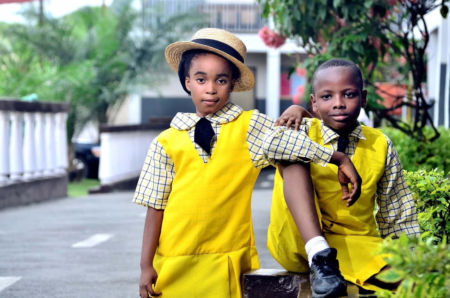 Two children in yellow school uniforms pose outdoors, with one wearing a straw hat and the other sitting with an arm on his friend's shoulder. There are trees and a building in the background.