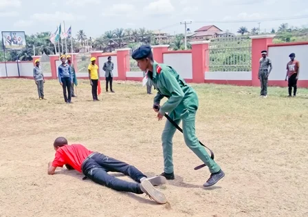 Person in a green uniform with a baton standing over someone lying on the ground wearing a red shirt and dark jeans, with several other people standing in the background.