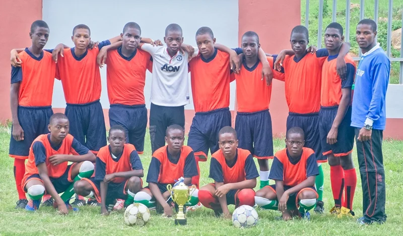 A youth soccer team of twelve boys and their coach posing outdoors on grass with a trophy and soccer balls, all wearing matching red and navy uniforms. Some players are kneeling in front, and others are standing behind.