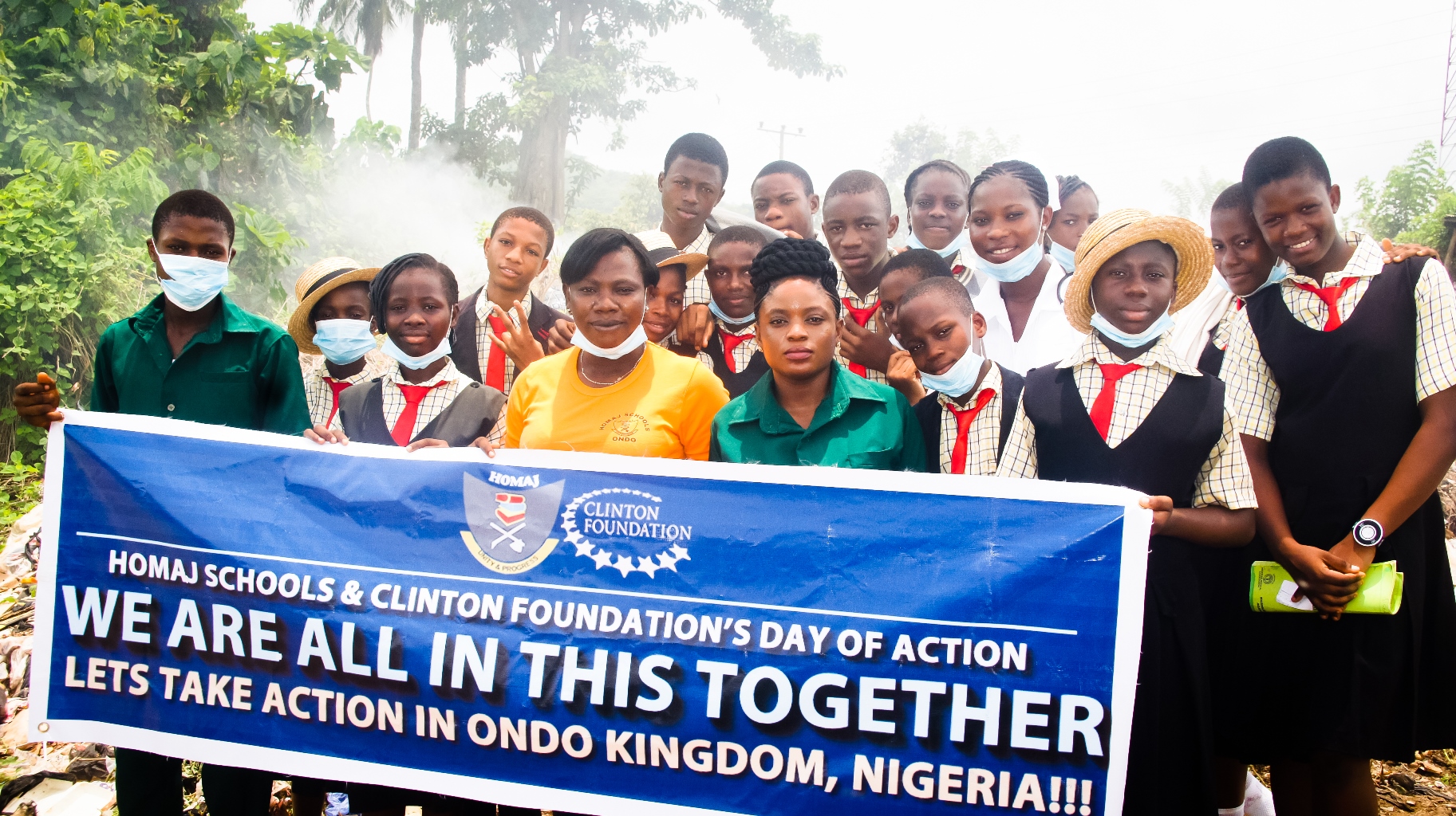 Group of school children and adults holding a banner for Homaj Schools and Clinton Foundation, celebrating Action Day in Ondo Kingdom, Nigeria, outdoors with greenery in the background.