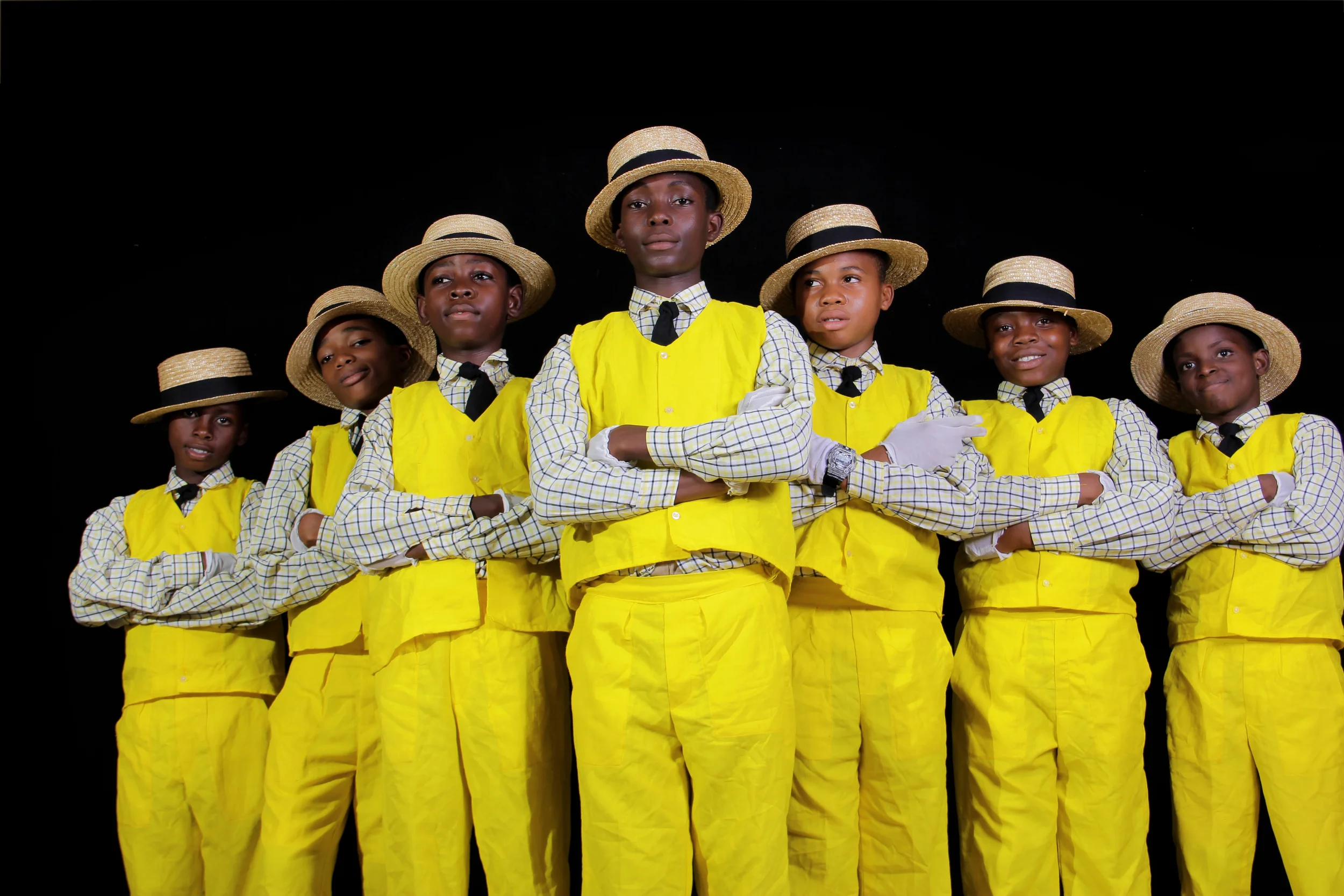 Seven young boys dressed in yellow vests, checkered shirts, black ties, and straw hats, standing with arms crossed against a black background.