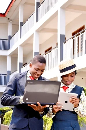 Two school children in uniform looking at a laptop and tablet outside a school building with balconies.