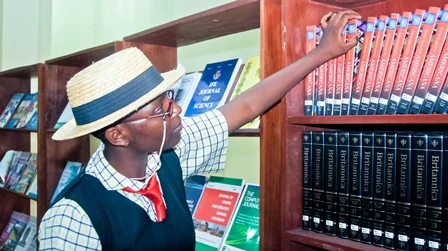 A person wearing a straw hat and glasses, reaching for a book on a library shelf, surrounded by books.