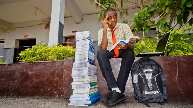 A young student in school uniform, sitting on a ledge outdoors, reading a book, with a large pile of papers, a backpack, and a laptop nearby.